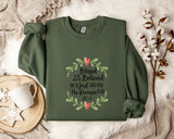 A green sweatshirt with a floral design and text printed on it, along with a mug and some white flowers on a bed.