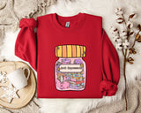 A red sweater with a graphic design of a jar containing various objects, including books and a teapot, is displayed on a white fur-covered surface.