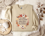 A beige sweatshirt with a floral wreath design and a quote printed on it, along with a mug, a branch of cotton bolls, and a snowflake ornament.