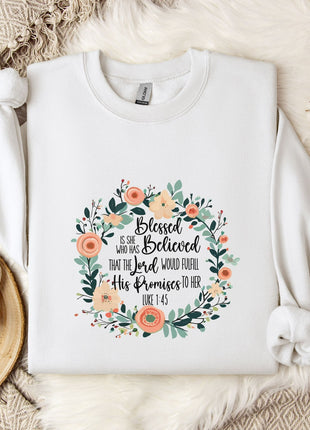 A white sweatshirt with a floral design and text printed on it, placed on a bed with a white blanket and a white mug.
