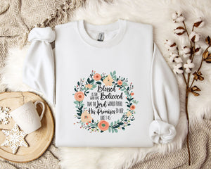 A white sweatshirt with a floral design and text printed on it, placed on a bed with a white blanket and a white mug.