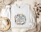 A white sweatshirt with a floral design and text printed on it, placed on a bed with a white blanket and a white mug.