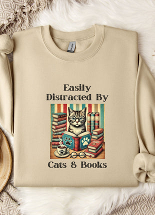 a beige sweatshirt with a printed design of a cat surrounded by books and a cup of coffee, along with the text "Easily Distracted By Cats & Books".