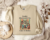 a beige sweatshirt with a printed design of a cat surrounded by books and a cup of coffee, along with the text "Easily Distracted By Cats & Books".