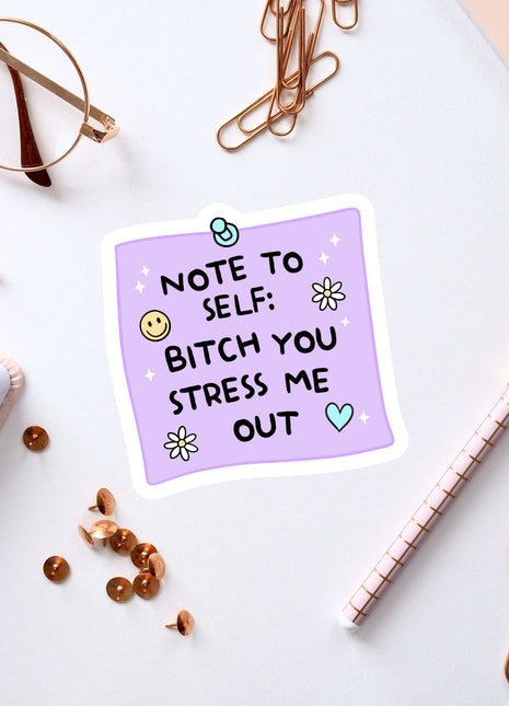 a purple sticker with a humorous message, surrounded by various office supplies and accessories such as a pen, a pair of glasses, and a notebook.