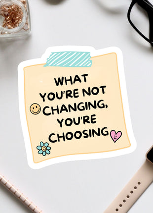 a sticker with a message that reads "What you're not changing, you're choosing", accompanied by a heart and a flower. The sticker is placed on a white surface, surrounded by various office supplies such as a pair of glasses, a pen, and a notebook.