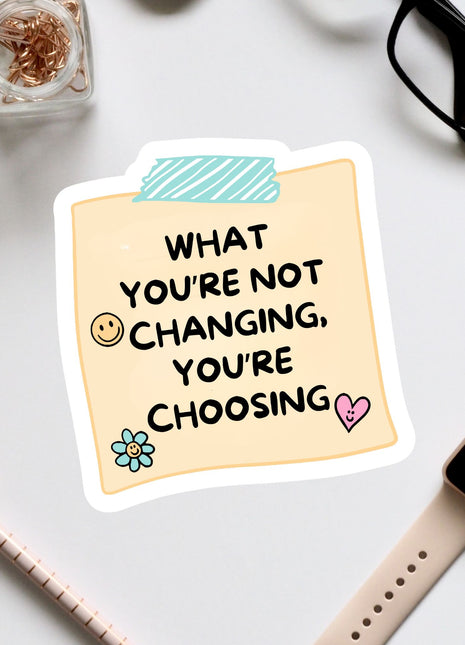 a sticker with a message that reads "What you're not changing, you're choosing", accompanied by a heart and a flower. The sticker is placed on a white surface, surrounded by various office supplies such as a pair of glasses, a pen, and a notebook.