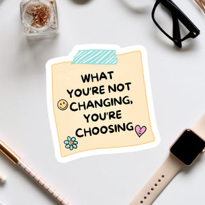 a sticker with a message that reads "What you're not changing, you're choosing", accompanied by a heart and a flower. The sticker is placed on a white surface, surrounded by various office supplies such as a pair of glasses, a pen, and a notebook.