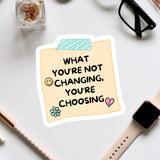 a sticker with a message that reads "What you're not changing, you're choosing", accompanied by a heart and a flower. The sticker is placed on a white surface, surrounded by various office supplies such as a pair of glasses, a pen, and a notebook.