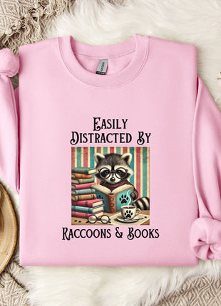 A pink sweatshirt with a graphic design of a raccoon reading books, accompanied by the text "Easily Distracted By Raccoons & Books".