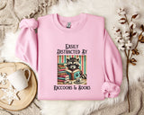 A pink sweatshirt with a graphic design of a raccoon reading books, accompanied by the text "Easily Distracted By Raccoons & Books".