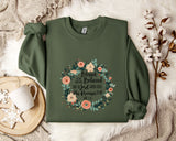 A green sweatshirt with a floral wreath design and text printed on it, placed on a white fur rug.