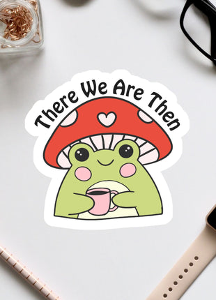 a sticker of a frog wearing a mushroom hat and holding a cup, surrounded by various office supplies such as a pair of glasses, a watch, and a pen.
