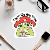 a sticker of a frog wearing a mushroom hat and holding a cup, surrounded by various office supplies such as a pair of glasses, a watch, and a pen.