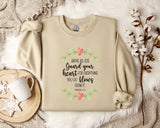 a beige sweatshirt with a printed Bible verse and a floral design, along with a mug, a white mug with a heart design, and some white flowers on a white fur-covered surface.