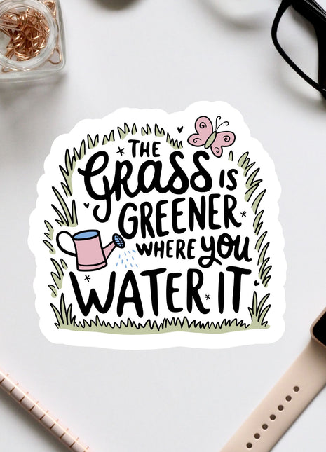 a sticker with a hand-drawn illustration of a watering can and a butterfly, surrounded by text that reads "The grass is greener where you water it".