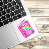 A sticker with a colorful book illustration is placed on a laptop keyboard, with a wooden surface in the background.
