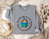 a gray sweatshirt with a graphic design depicting a fire-breathing trash can surrounded by flames and smoke. The sweatshirt is placed on a bed with a white blanket and a white mug.