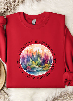 A red sweater with a circular design featuring a forest scene and the text "AND INTO THE FOREST I GO TO TOLOSSO" is displayed on a bed with a white blanket and a white mug.