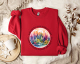 A red sweater with a circular design featuring a forest scene and the text "AND INTO THE FOREST I GO TO TOLOSSO" is displayed on a bed with a white blanket and a white mug.