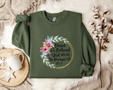 A green sweatshirt with a floral design and text printed on it, along with a mug and some white flowers on a bed.