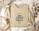 A beige sweatshirt with a printed Bible verse and a floral design, along with a mug, a white mug with a heart design, and some white cotton balls, all placed on a white fur rug.