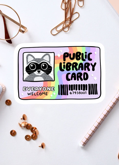 a sticker with a raccoon and the words "Public Library Card" on a white background. There are also various office supplies, such as a pen, a pair of glasses, and a notebook, scattered around the sticker.