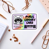 a sticker with a raccoon and the words "Public Library Card" on a white background. There are also various office supplies, such as a pen, a pair of glasses, and a notebook, scattered around the sticker.