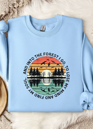 A light blue sweatshirt with a graphic design depicting a sunset over a forest, along with the text "And into the forest I go, and I lose my soul" printed on it. The sweatshirt is laid out on a bed with a white blanket and a white mug, and there are some white flowers nearby.