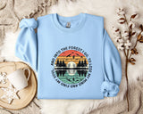 A light blue sweatshirt with a graphic design depicting a sunset over a forest, along with the text "And into the forest I go, and I lose my soul" printed on it. The sweatshirt is laid out on a bed with a white blanket and a white mug, and there are some white flowers nearby.