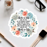 a sticker with a floral design and text, surrounded by various office supplies such as a watch, pen, and glasses.