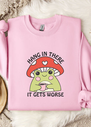 A pink sweatshirt with a mushroom graphic and the text "HANG IN THERE IT GETS WORSE" printed on it, along with a mug and some white flowers on a bed.