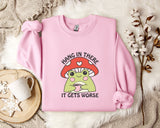 A pink sweatshirt with a mushroom graphic and the text "HANG IN THERE IT GETS WORSE" printed on it, along with a mug and some white flowers on a bed.