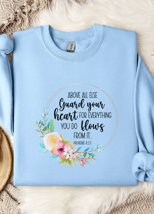 A light blue sweatshirt with a floral design and text printed on it, placed on a bed with a white blanket and a white mug.