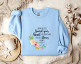 A light blue sweatshirt with a floral design and text printed on it, placed on a bed with a white blanket and a white mug.