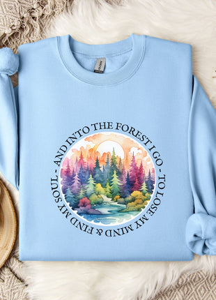 A light blue sweatshirt with a colorful forest scene printed on it, along with a mug and some flowers, all laid out on a bed with a white blanket.