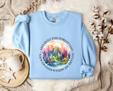 A light blue sweatshirt with a colorful forest scene printed on it, along with a mug and some flowers, all laid out on a bed with a white blanket.
