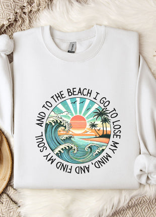 A white sweatshirt with a graphic design depicting a beach scene, along with the text "And to the beach I go, I lose my mind" printed on it. The sweatshirt is laid out on a bed with a floral patterned blanket, and there is a mug and a branch of cotton balls nearby.