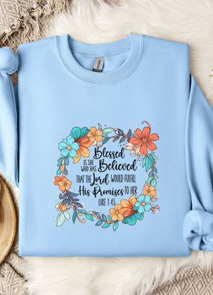 A light blue sweatshirt with a floral design and text printed on it, placed on a white fur rug. Next to the sweatshirt are a white mug, a white teapot, and some white flowers.