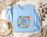 A light blue sweatshirt with a floral design and text printed on it, placed on a white fur rug. Next to the sweatshirt are a white mug, a white teapot, and some white flowers.