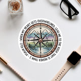 a compass with a mountain landscape and a quote around it, along with some office supplies and a pair of glasses on a white surface.