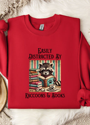 A red sweatshirt with a graphic of a raccoon reading books, accompanied by the text "Easily Distracted By Raccoons & Books".