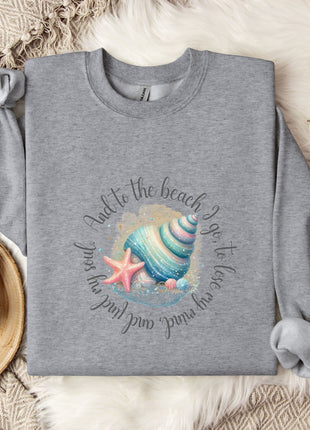 a gray sweatshirt with a seashell and starfish design, along with a mug and a branch of cotton flowers, all placed on a beige blanket.