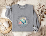 a gray sweatshirt with a seashell and starfish design, along with a mug and a branch of cotton flowers, all placed on a beige blanket.