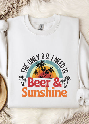 A white sweatshirt with a graphic design that says "The only B.S. I need is Beer & Sunshine" printed on it, along with a palm tree and sunset. The sweatshirt is laid out on a bed with a floral blanket, and there is a mug and some cotton balls nearby.