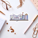 a sticker with a dragon and various objects, such as books, a pen, and a pair of glasses, arranged on a white surface.