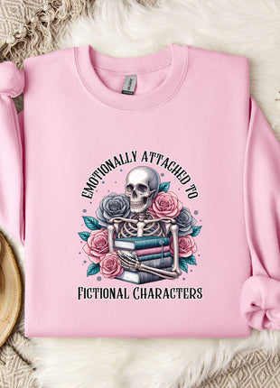 A pink sweatshirt with a graphic design of a skeleton surrounded by flowers and books, along with the text "Emotionally Attached to Fictional Characters".