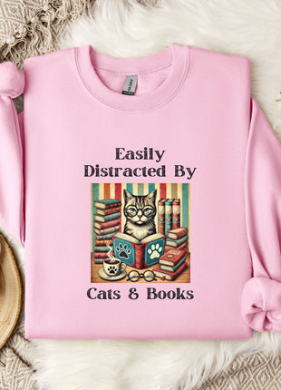 A pink sweatshirt with a cat illustration and the text "Easily Distracted By Cats & Books" printed on it, along with a mug and a branch of cotton bolls.