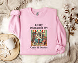 A pink sweatshirt with a cat illustration and the text "Easily Distracted By Cats & Books" printed on it, along with a mug and a branch of cotton bolls.
