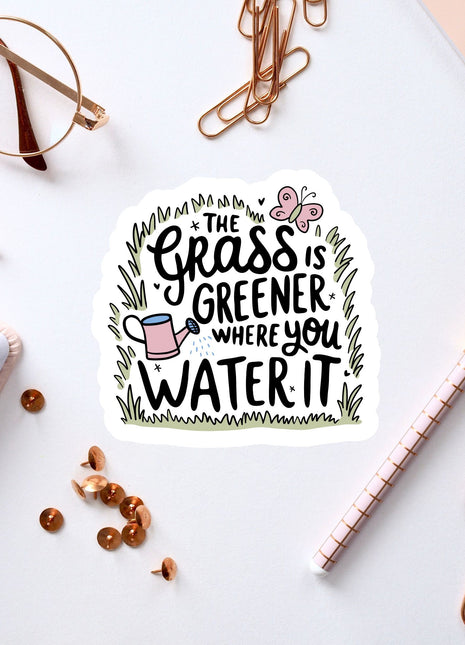 a sticker with a hand-drawn illustration of a watering can surrounded by grass, along with the text "The grass is greener where you water it".